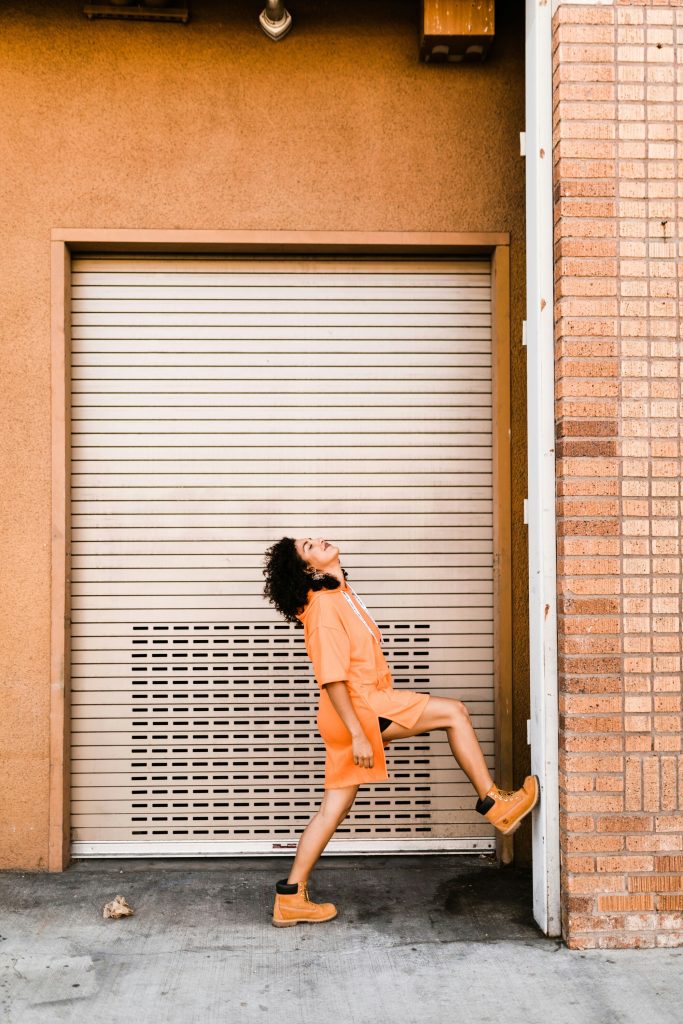 Mujer con conjunto deportivo naranja y botas urbanas, posando frente a una puerta metálica en un entorno urbano.